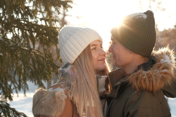 A man and a woman in winter clothes looking at each other in the light of the sun in a winter forest