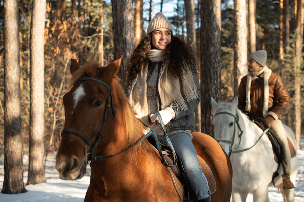 A dark-haired woman in a white hat and warm outerwear riding a brown stallion in a snowy forest