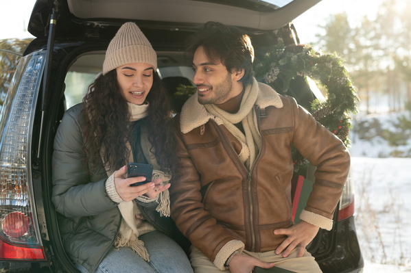 A couple dressed in warm clothes surfing the Internet on the phone while sitting in the car trunk on a winter date