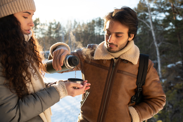 A dark-haired man pouring a hot drink from a thermos to his girlfriend on a date in the winter forest