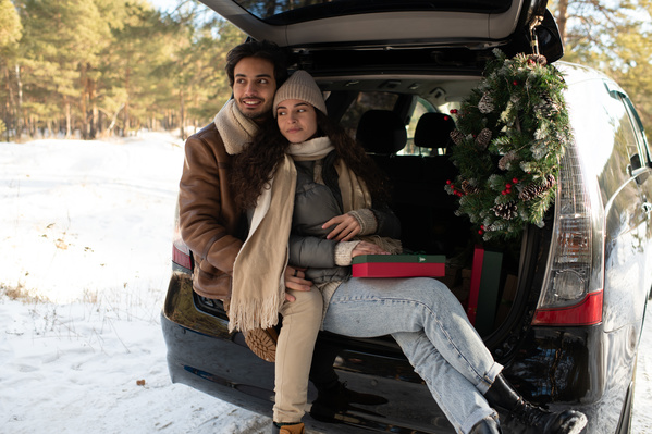 A man and his girlfriend dressed warmly with Christmas gifts in bright boxes posing in the car trunk in a winter forest