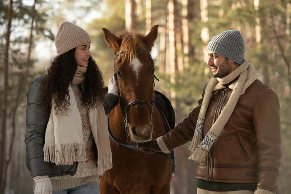 A man in warm clothes and his dark-haired girlfriend walking through the forest with a brown stallion