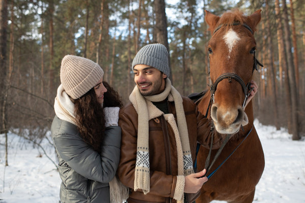 A romantic couple dressed warmly walking with a brown horse in a winter forest
