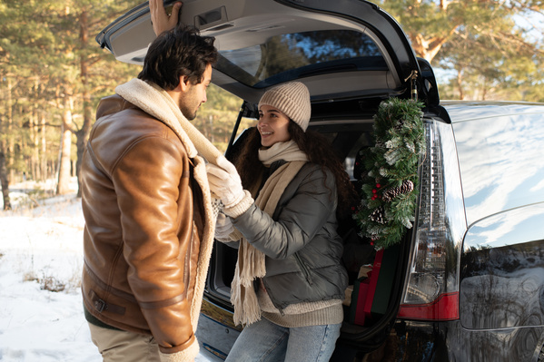 A dark-haired woman in warm clothes sitting in the car trunk and her boyfriend in a winter forest