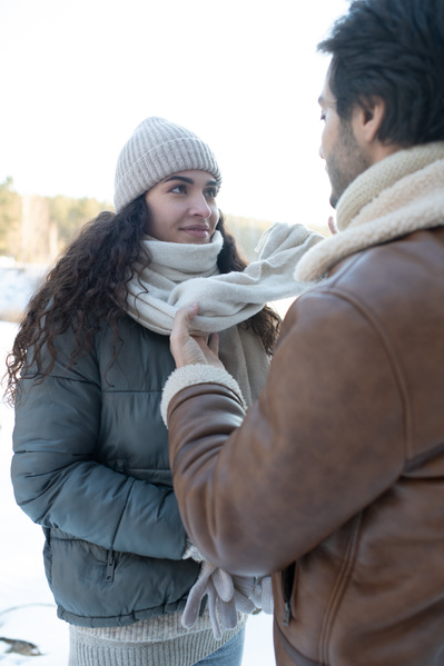 A man in a warm sheepskin coat tying a scarf to his dark-haired girlfriend on a winter walk