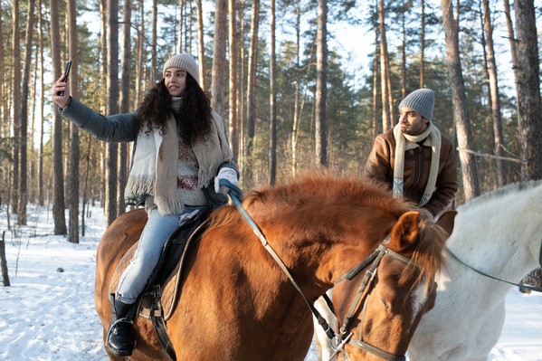 A dark-haired woman taking a selfie with her boyfriend on a horse ride in the winter forest