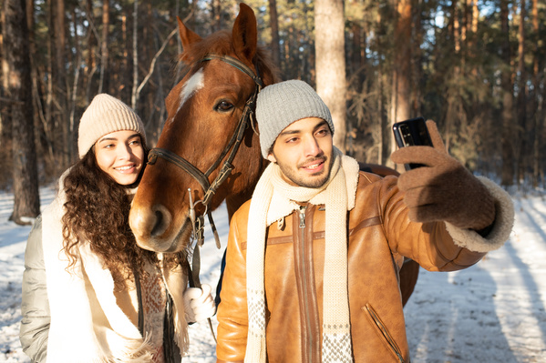A man in warm clothes taking a selfie with his dark-haired girlfriend on a horse walking in the winter forest