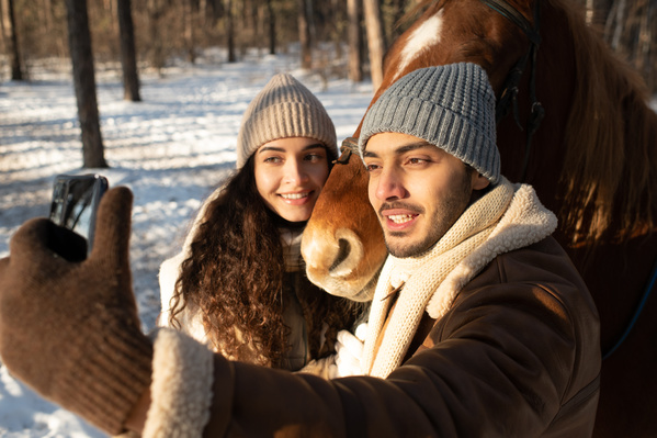 A guy in warm clothes taking a selfie with his girlfriend on a horse ride in the winter forest