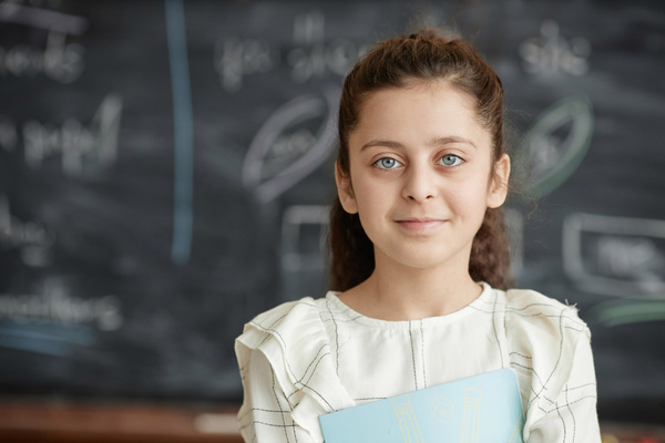 In this image a young girl is standing in front of a chalkboard in a classroom setting.