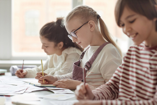 In this image there are three young girls sitting at a desk each holding a pencil and engrossed in drawing or coloring.