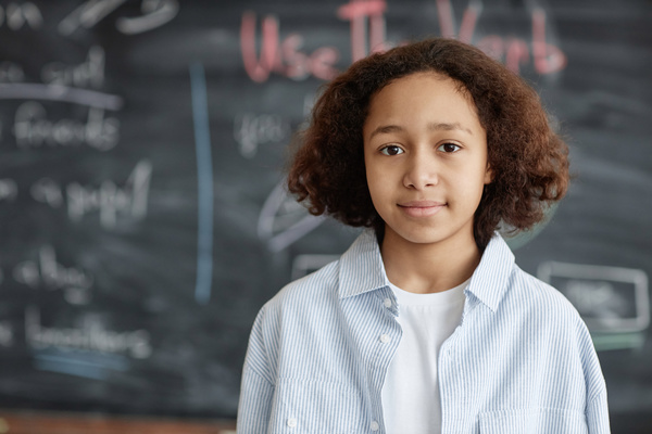 In this image a young girl is standing in front of a chalkboard smiling at the camera.