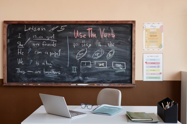 The image depicts a classroom with a chalkboard a desk and a laptop.