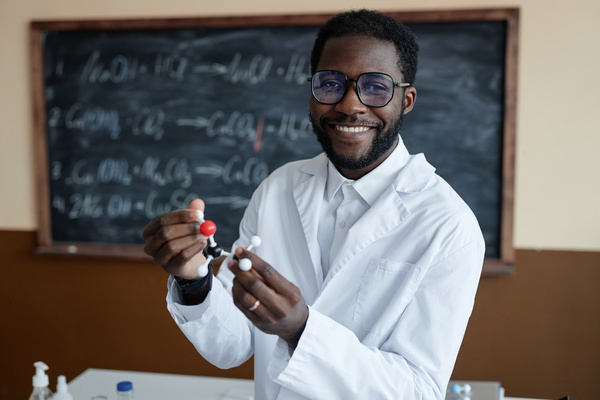 In the image a smiling man wearing a white lab coat and glasses is standing in front of a chalkboard.