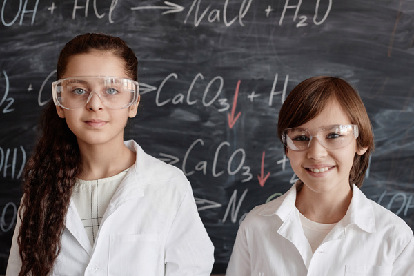 In this image two young girls are standing in front of a chalkboard both wearing white lab coats and goggles.