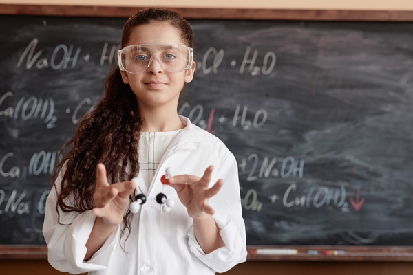 In this image a young girl is standing in front of a chalkboard wearing a lab coat and goggles.