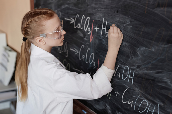 In this image a young girl wearing a lab coat and goggles is writing on a chalkboard.