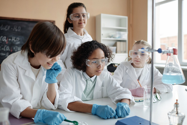 The image depicts a group of four young children wearing lab coats and goggles working together on a science experiment in a classroom or laboratory setting.