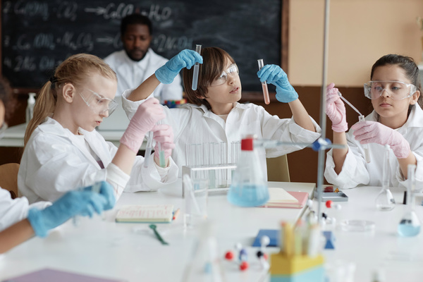 The image depicts a group of young children wearing white lab coats and gloves engrossed in a science experiment.