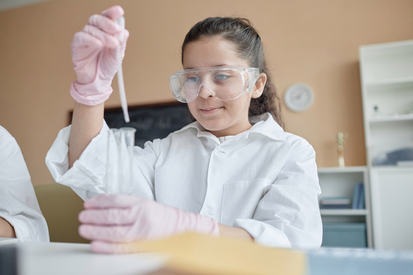 In the image a young girl wearing a lab coat and goggles is performing a science experiment in a laboratory setting.