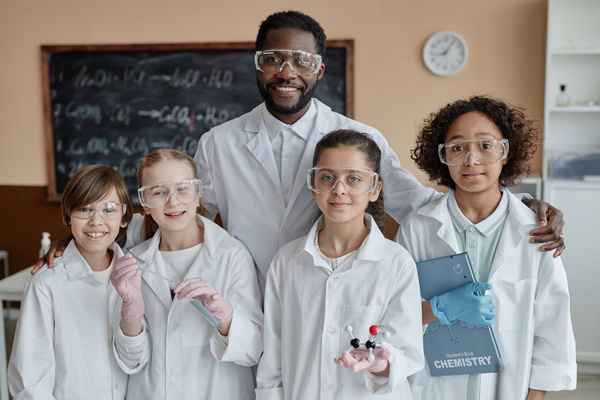The image depicts a group of five young children dressed in white lab coats and goggles standing together in a classroom or laboratory setting.