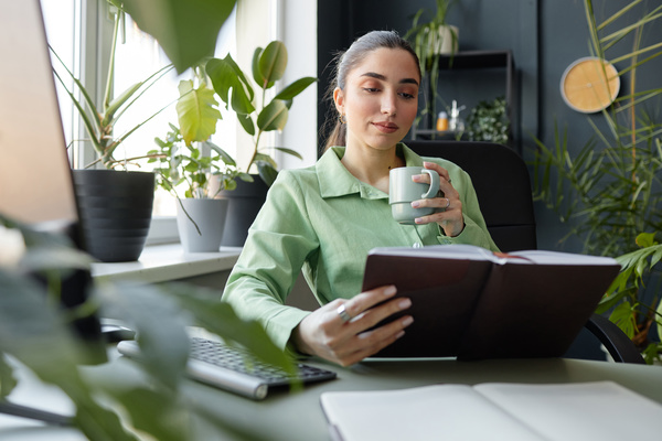 In the image a woman is sitting at a desk in a green shirt holding a cup of coffee and reading a book.