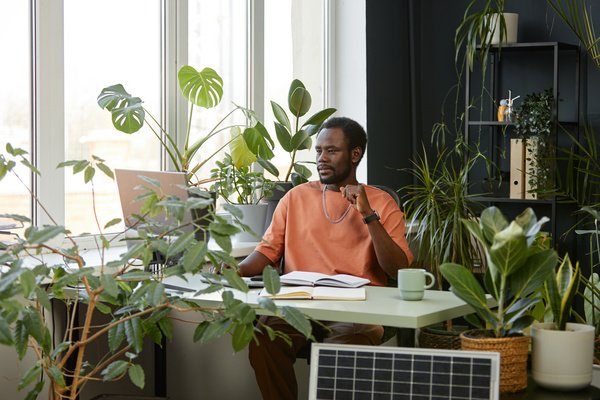 In this image a man is sitting at a desk surrounded by a variety of potted plants.