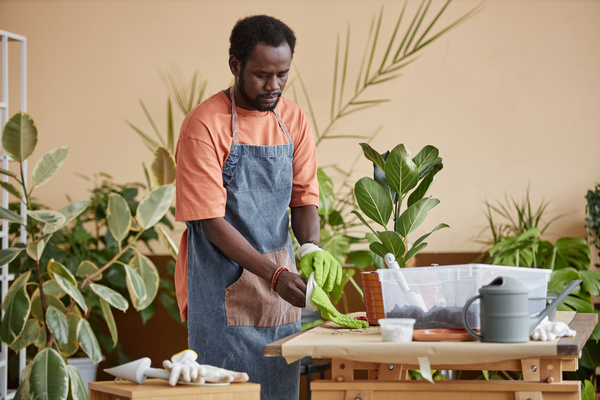 In the image a man wearing an apron is working on a wooden table surrounded by various potted plants.