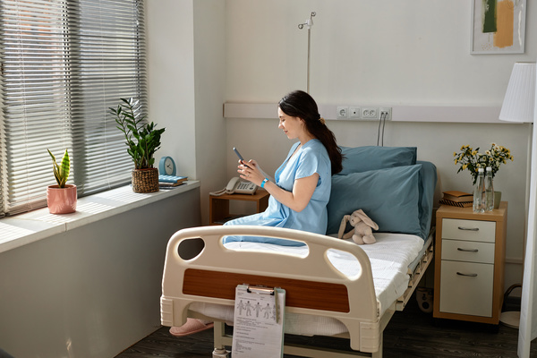 The image depicts a woman sitting in a hospital bed holding a tablet device.