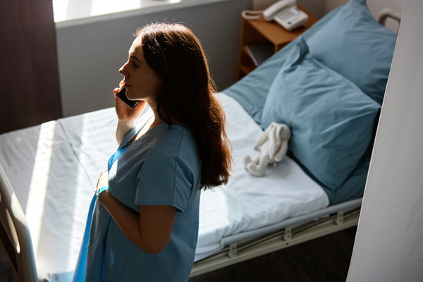In this image a woman is standing in front of a hospital bed holding a cell phone to her ear.