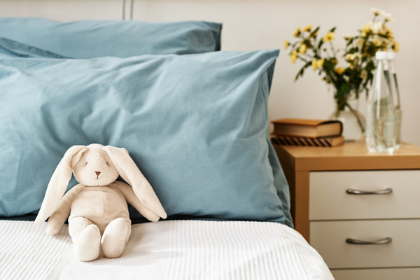 In the image there is a small stuffed bunny rabbit sitting on a bed positioned between two blue pillows.