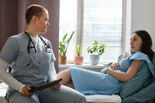 In this image a female nurse is sitting on a bed next to a pregnant woman.