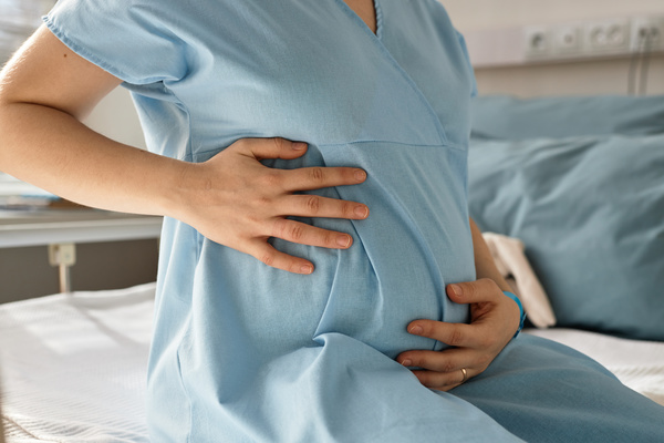 In this image a pregnant woman is sitting on a bed with her hands on her stomach.