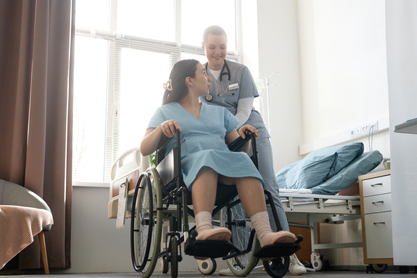 In this image a woman in a wheelchair is being assisted by a nurse in a hospital room.