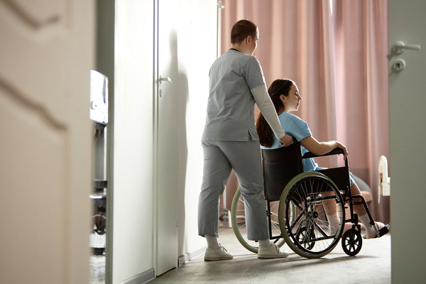 In this image a nurse is pushing a woman in a wheelchair down a hallway.