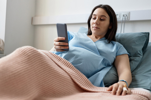 In this image a pregnant woman is lying in a hospital bed holding a smartphone in her hand.