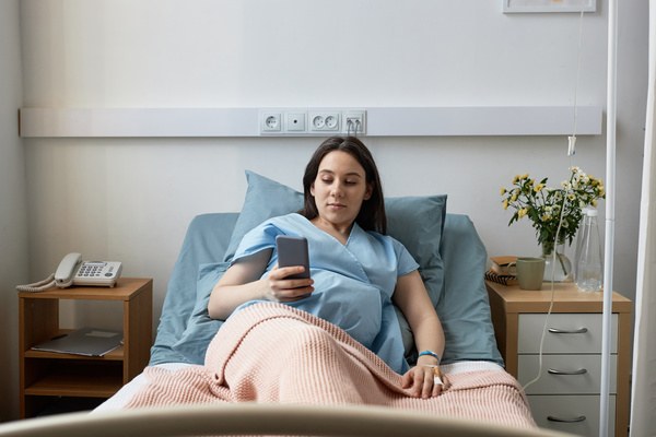 In this image a pregnant woman is lying in a hospital bed holding a cell phone in her hand.