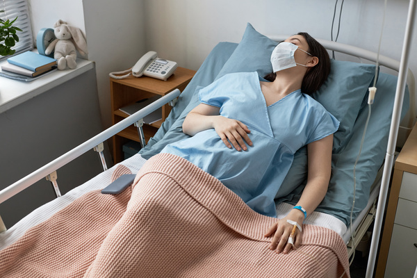 In this image a pregnant woman is lying in a hospital bed wearing a blue hospital gown and a face mask.