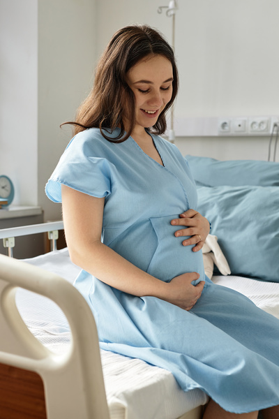 In this image a pregnant woman is sitting on a hospital bed wearing a blue dress.
