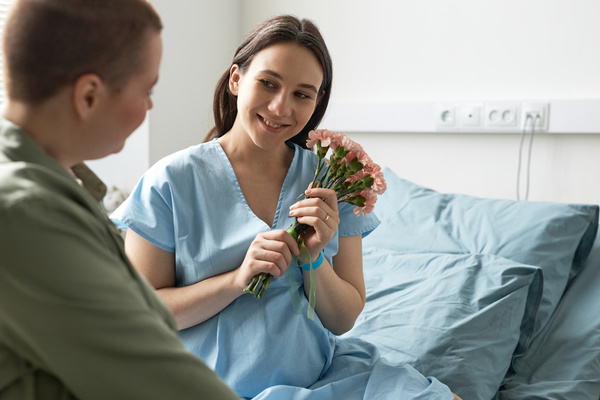 In this image a pregnant woman is sitting on a bed holding a bouquet of pink flowers.