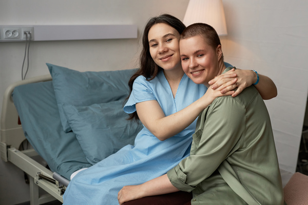 In this image two women are embracing each other while sitting on a hospital bed.