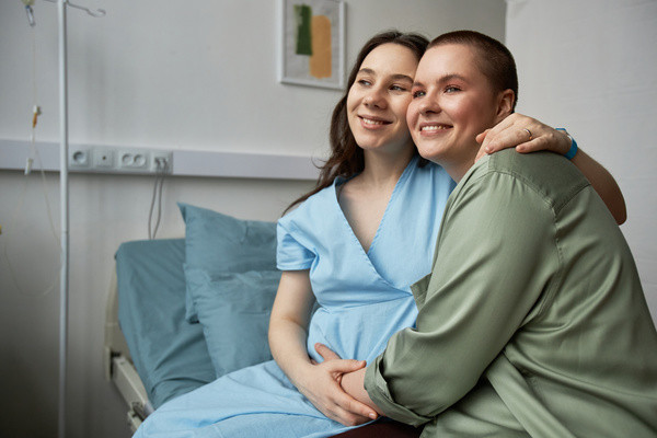 In this image two women are sitting on a hospital bed embracing and smiling at each other.