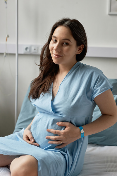 In this image a pregnant woman is sitting on a hospital bed smiling at the camera.