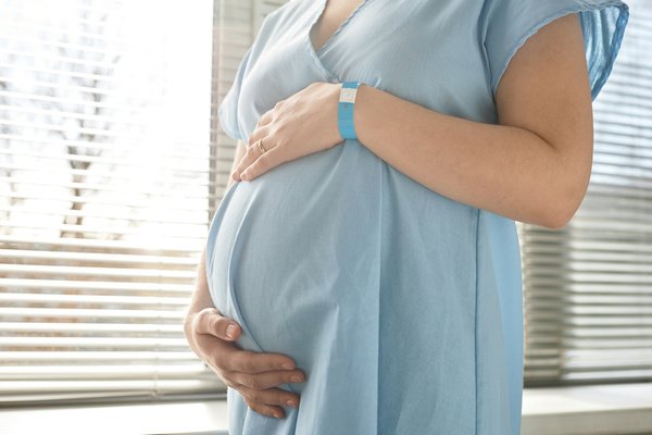 In this image a pregnant woman is standing in front of a window wearing a blue dress.