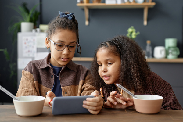 In this image two young girls are sitting at a dining table with a tablet in front of them.