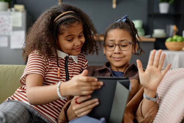 In this image two young girls are sitting on a couch together engrossed in an activity on a tablet.