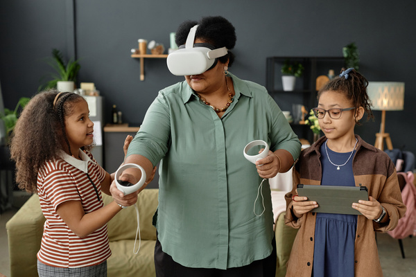 In the image a woman wearing a virtual reality headset is interacting with two young girls who are also wearing vr headsets.