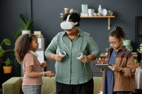 In the image a woman wearing virtual reality goggles is interacting with two young girls who are also wearing vr goggles.