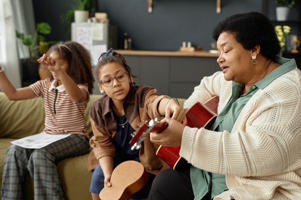 In this image a woman is sitting on a couch and playing a ukulele while two young girls watch her.