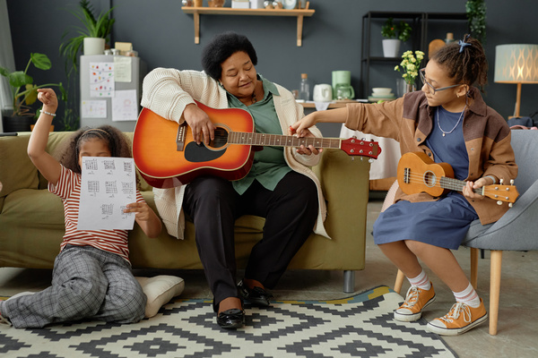 In the image an elderly woman is sitting on a couch playing a guitar surrounded by two young girls.