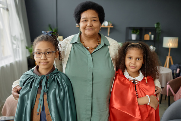 In the image a grandmother is posing with two young girls both wearing cape costumes.